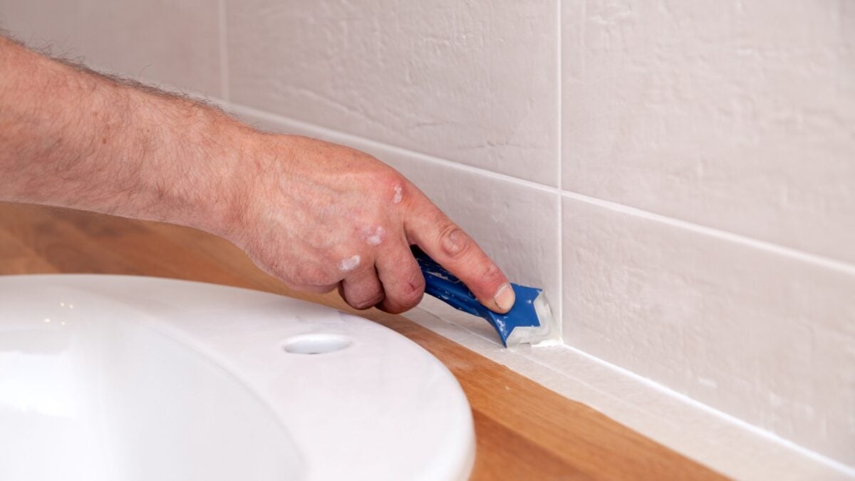 Closeup hand applying white sealant, joint compound, caulk to joint of bathroom tile using blue scraper.