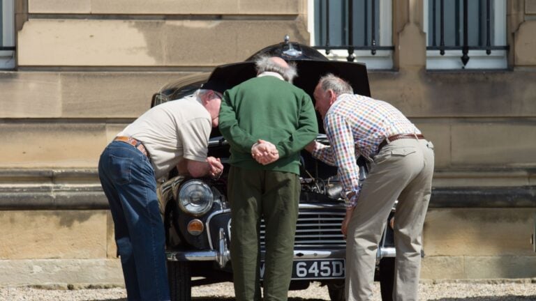 Three old men inspect the engine under the bonnet of a Morris Minor Classic Car.