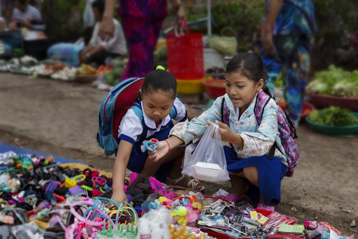 School Shopping Two young schoolgirls explore and purchase items at a lively outdoor market, filled with vibrant colors.