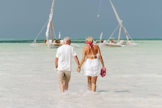 retire early An elderly couple holds hands while walking on the beach, with dhows in the background in Zanzibar.