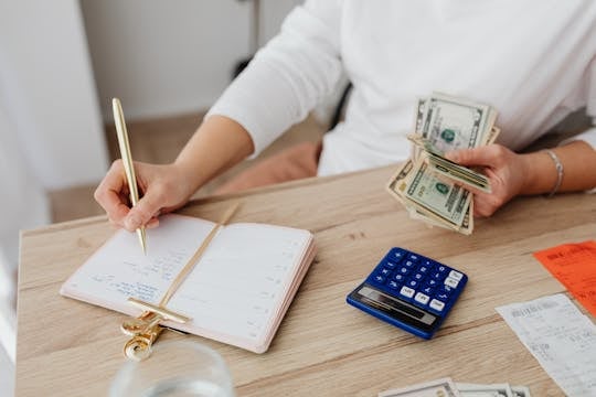 Inflation-Proof Your Budget Adult holding cash and writing in planner while using a calculator at home.