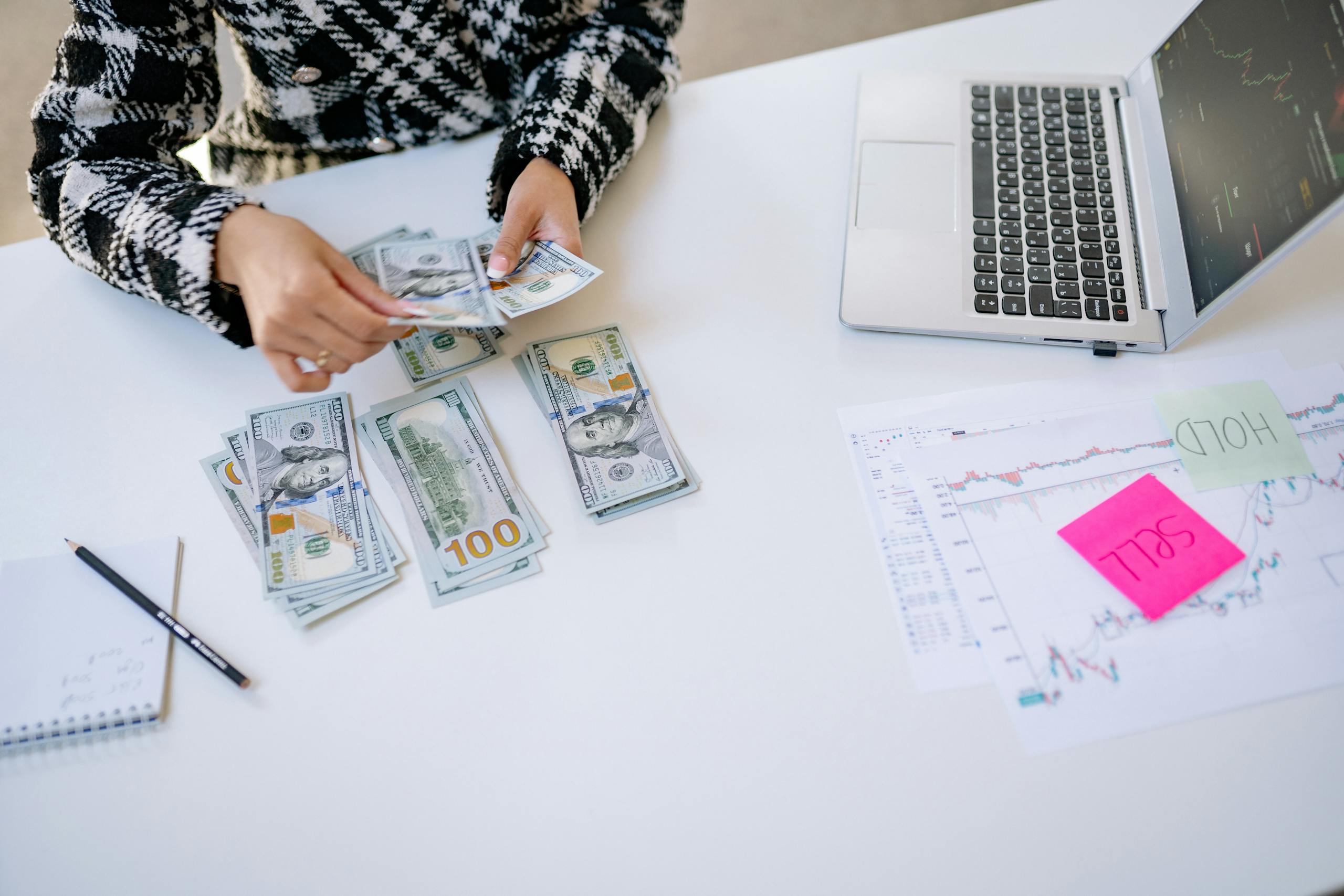 Negotiate Person counting cash next to laptop and stock market charts on a white table.