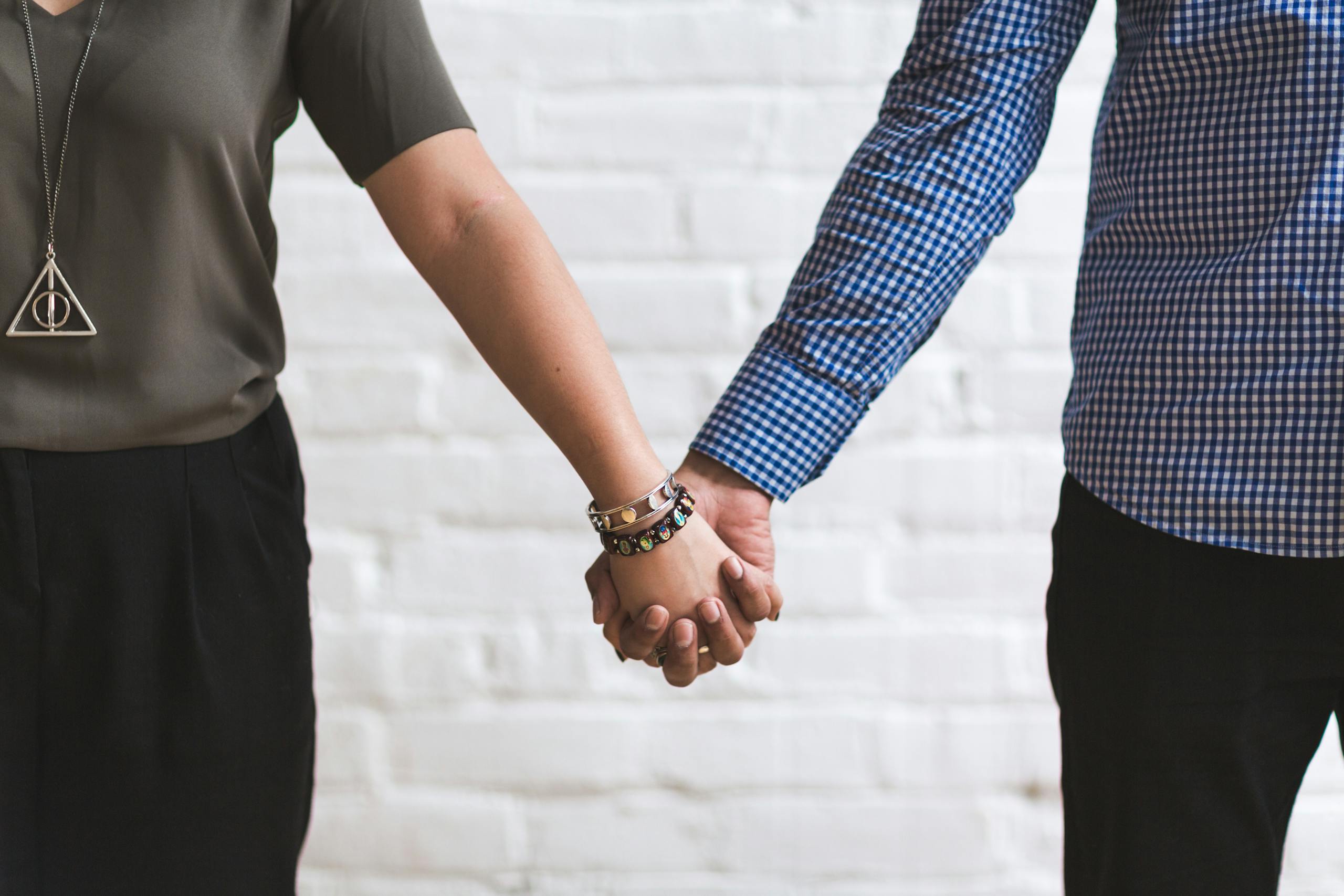 Financial Infidelity Close-up of a couple holding hands in casual fashion attire against a white brick wall.