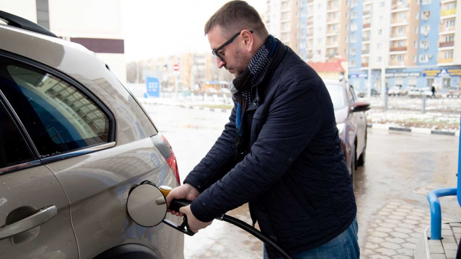 A man with a blue jacket and dark glasses at a gas station. He fills up the car. Petrol. Lifestile.