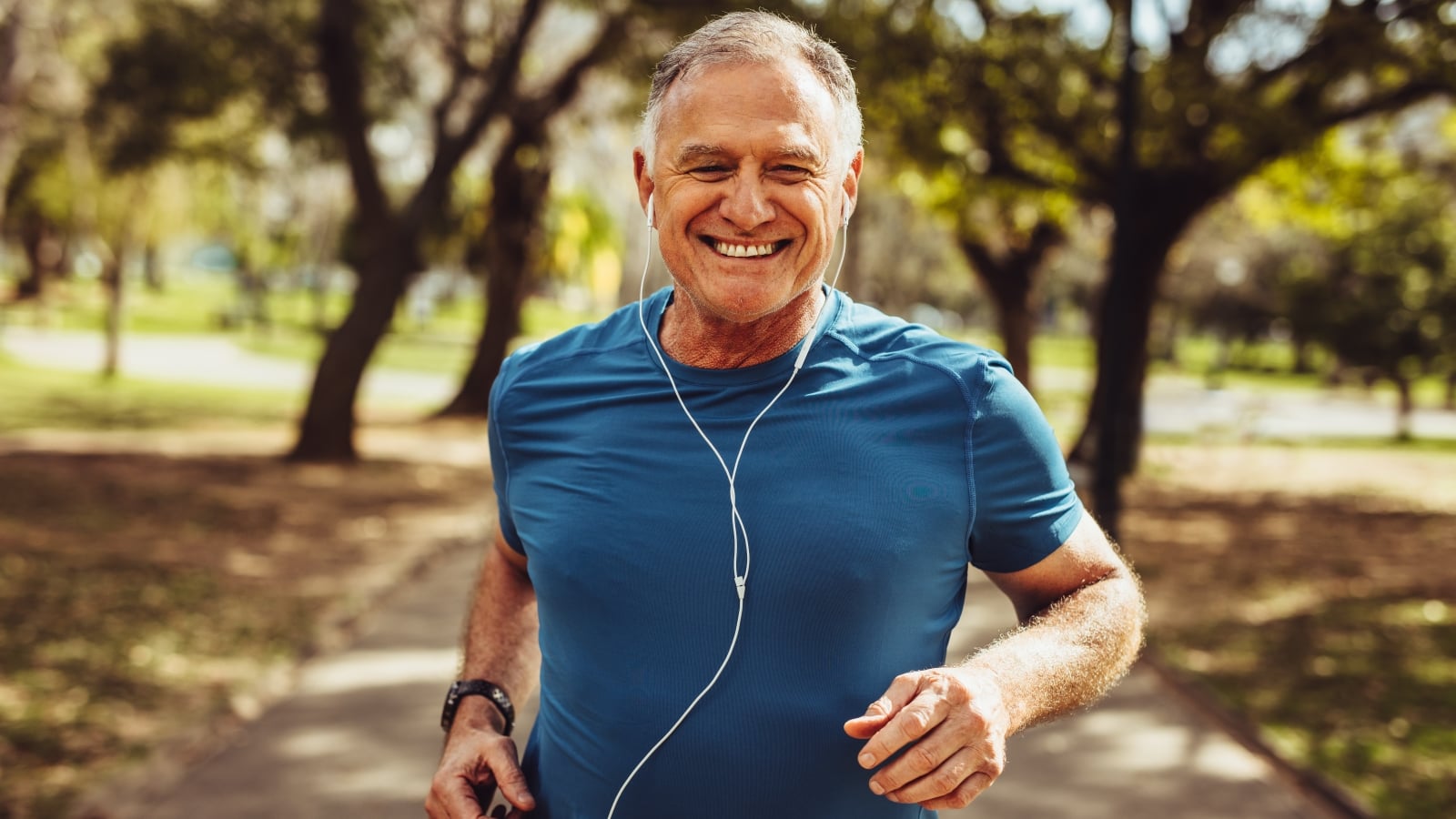 Portrait of a senior man in fitness wear running in a park. Close up of a smiling man running while listening to music using earphones