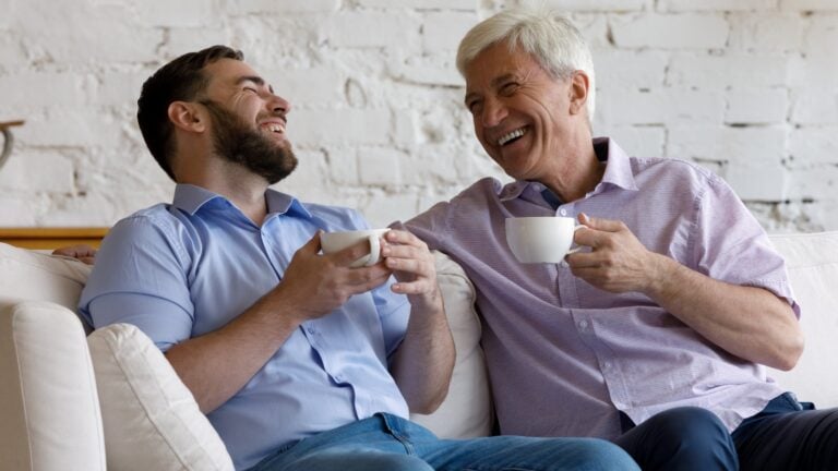 Happy adult son meeting with mature retired grey haired father, drinking hot tea, coffee, talking, laughing, enjoying family leisure time on weekend at home, keeping good healthy warm relationships